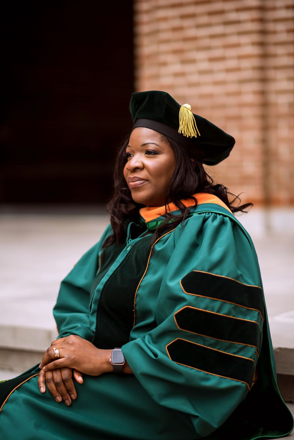 Graduate in emerald green academic regalia with black velvet trim poses for celebratory photos against brick wall. Rice University photos