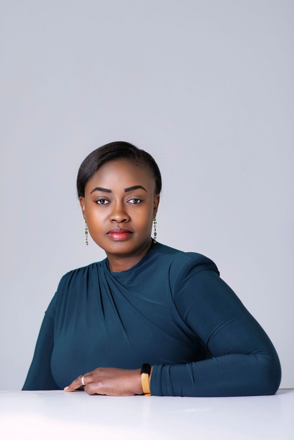 Professional portrait of someone in a dark teal long-sleeved top seated at a white desk against a light gray background.