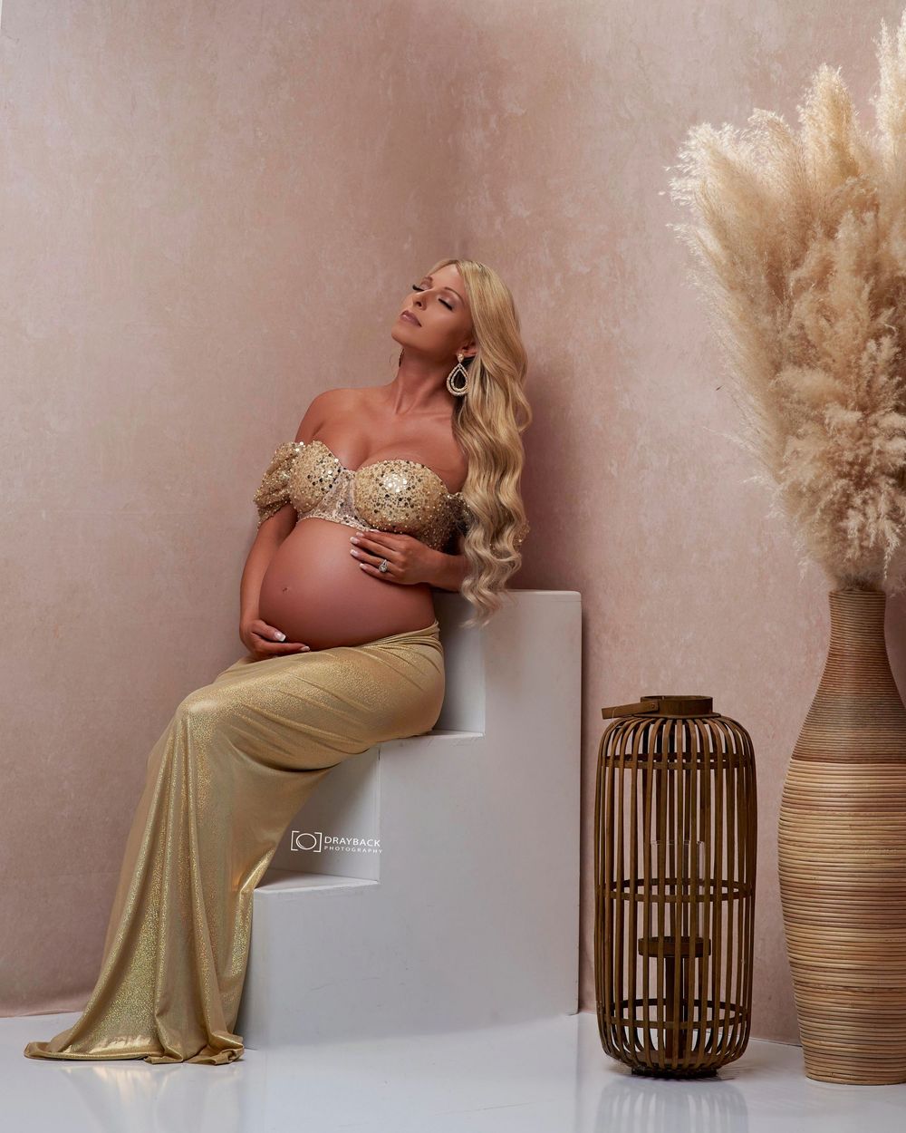 A maternity photoshoot series featuring a gold dress against a neutral backdrop with decorative pampas grass and wicker decor.