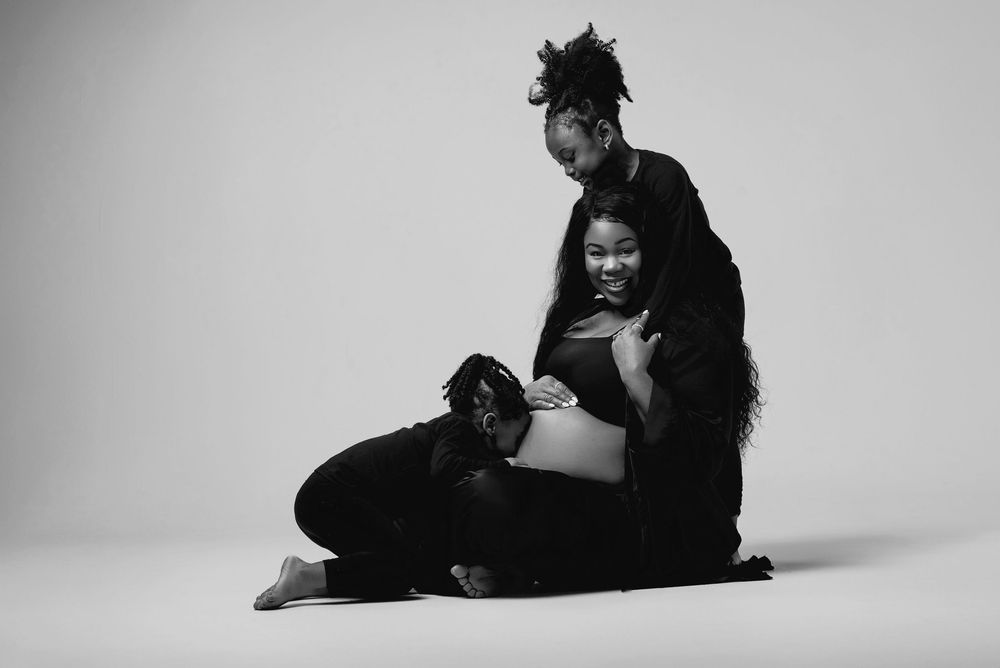 Artistic black and white maternity photo with figures in dark clothing against white background.