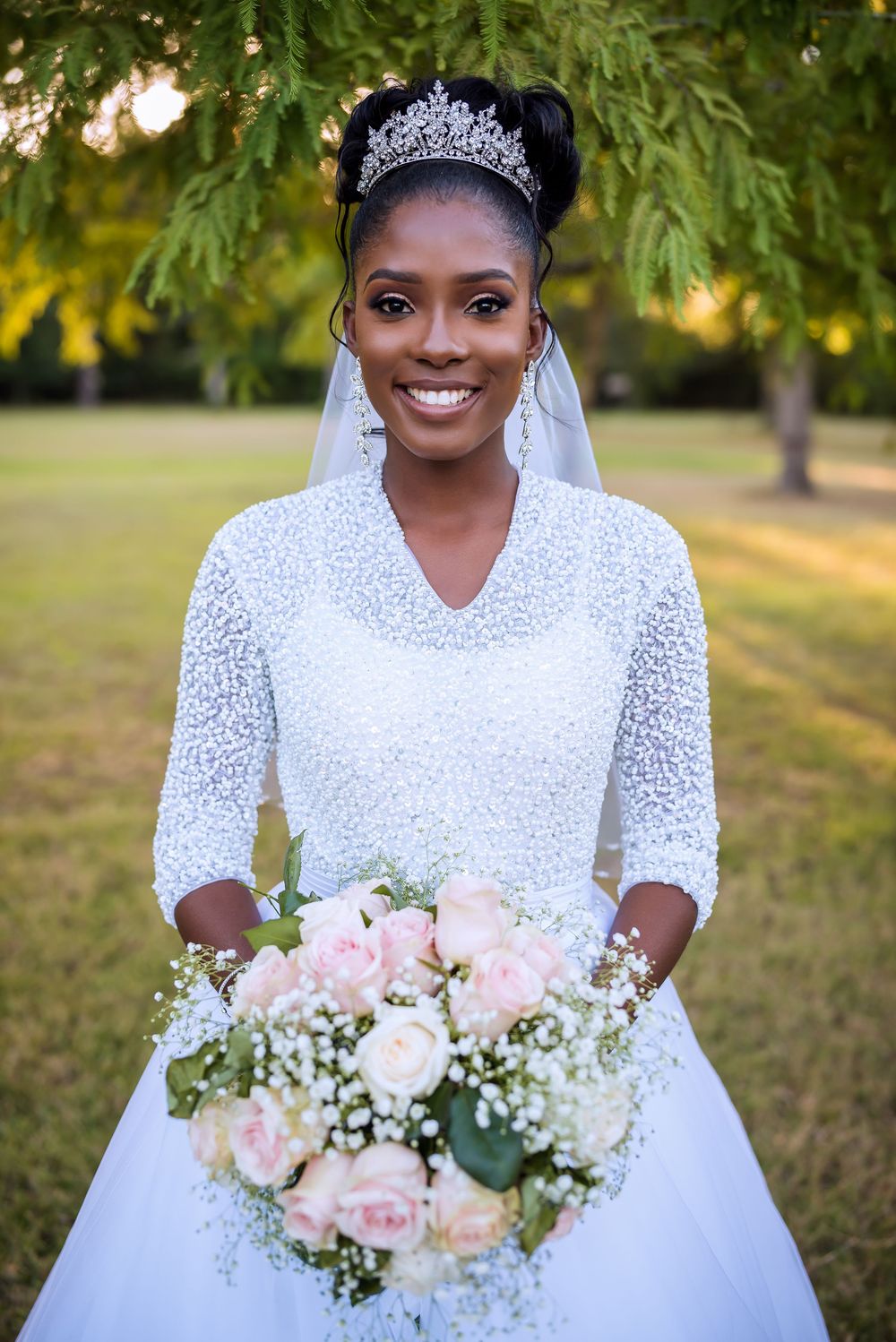 Beautiful bride in white lace gown with long sleeves poses with pink and white rose bouquet in outdoor garden setting.