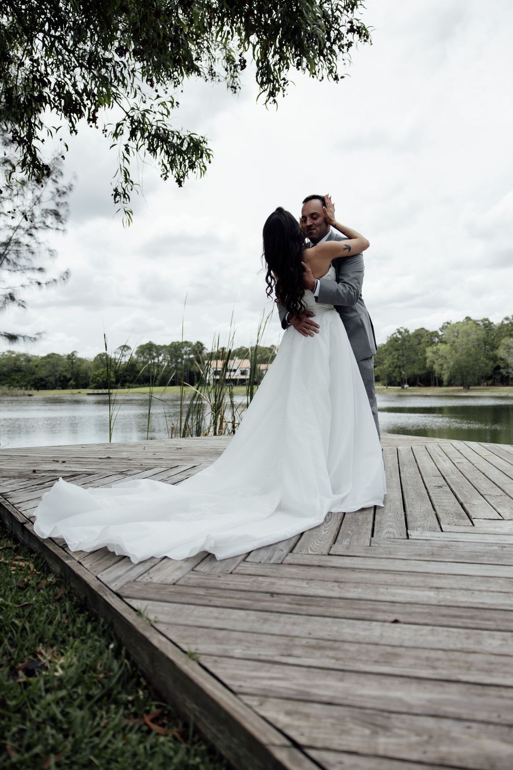 A couple embraces on a wooden dock overlooking a serene lake with overhanging trees.