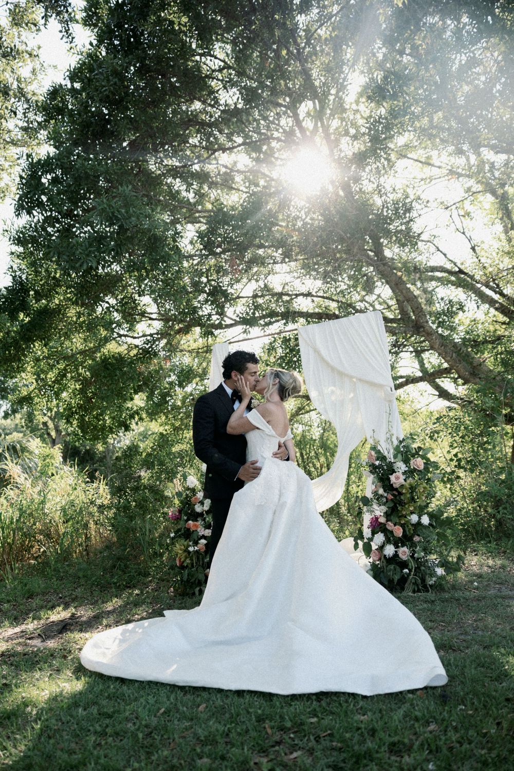 A couple in wedding attire shares a romantic moment under sunlit trees in a garden setting.