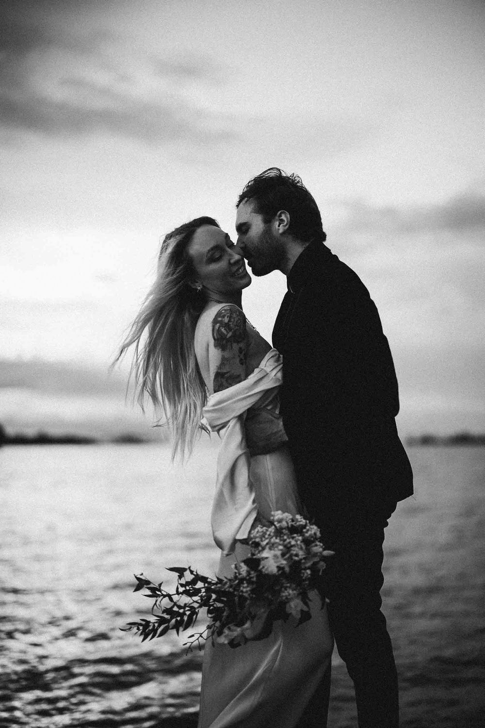 A romantic black and white photo sequence of a couple embracing by the water at sunset with a floral bouquet.