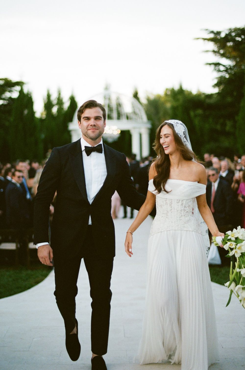 Newlyweds walk together on a path surrounded by wedding guests during an outdoor evening celebration.