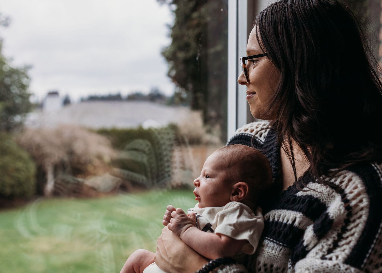 A person wearing a cozy knit sweater holds a small child while looking out a window into nature.