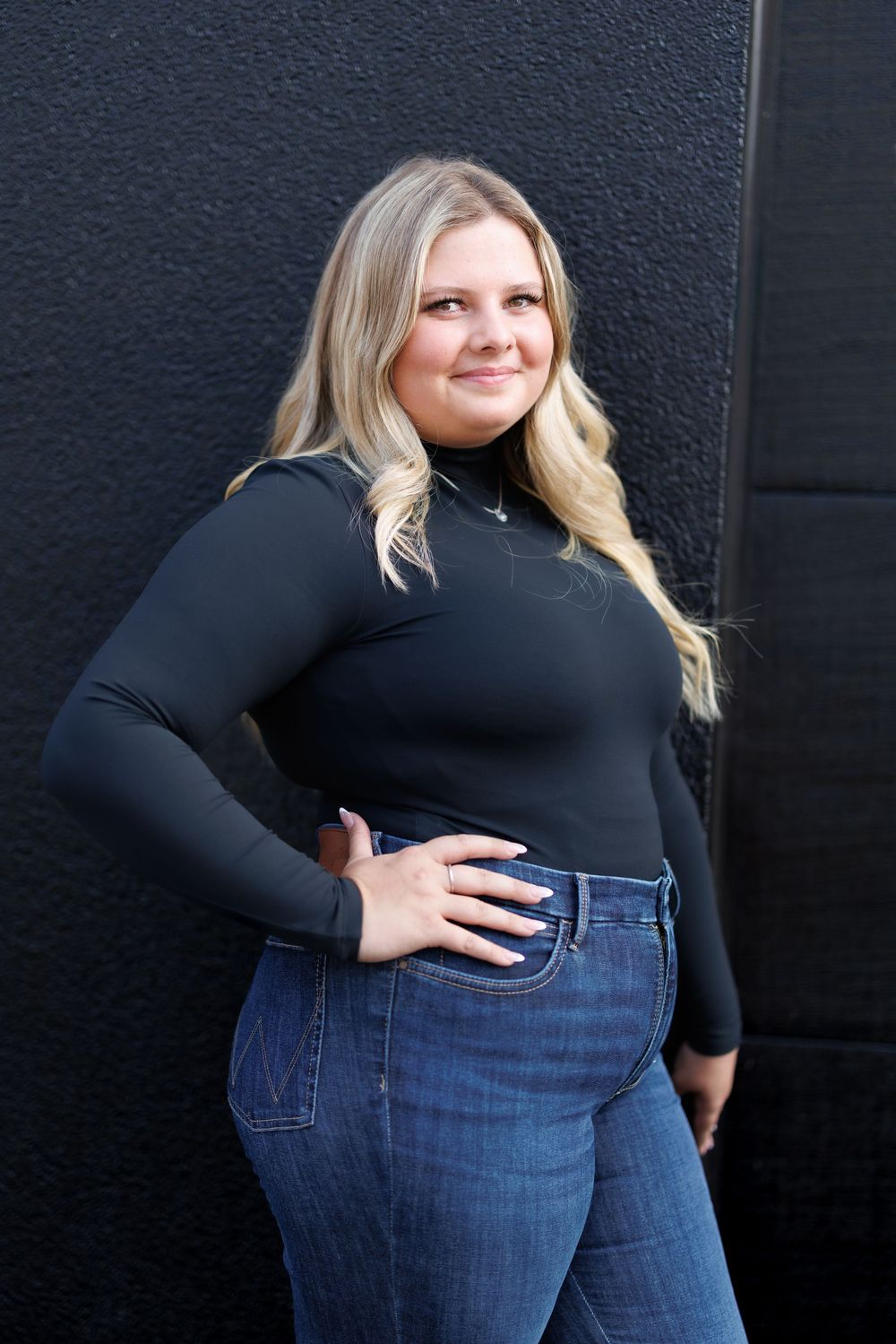Fashion portrait against black wall wearing black turtleneck and blue jeans.