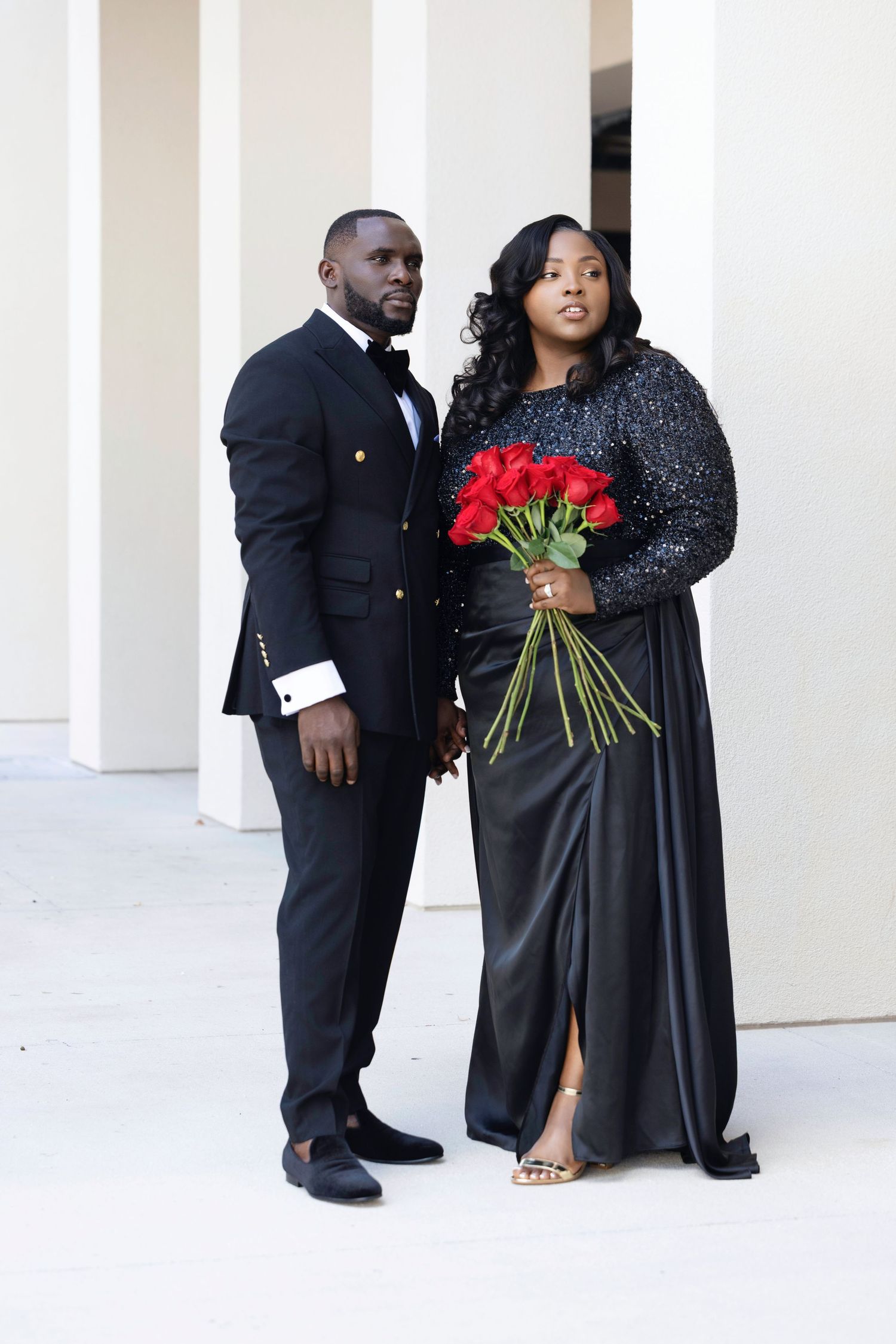 A romantic couple in formal black attire poses for engagement photos with red roses against a white column backdrop.