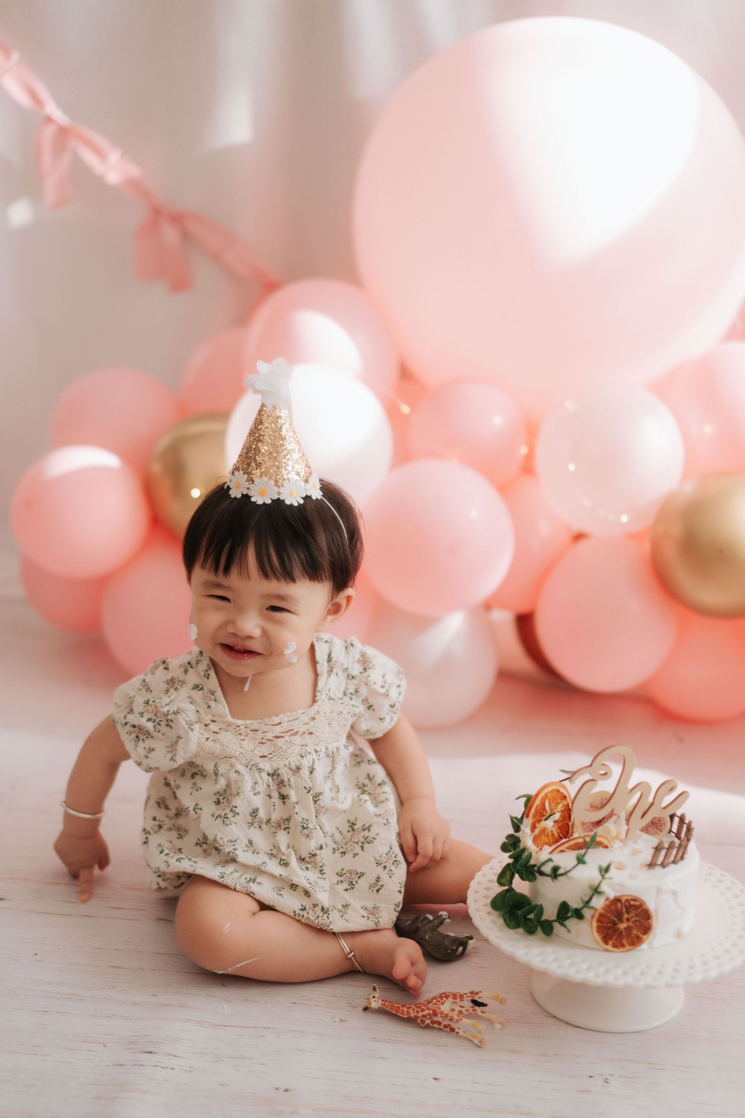 A cake smash photo session featuring a baby in a white dress surrounded by balloons and decorative elements.