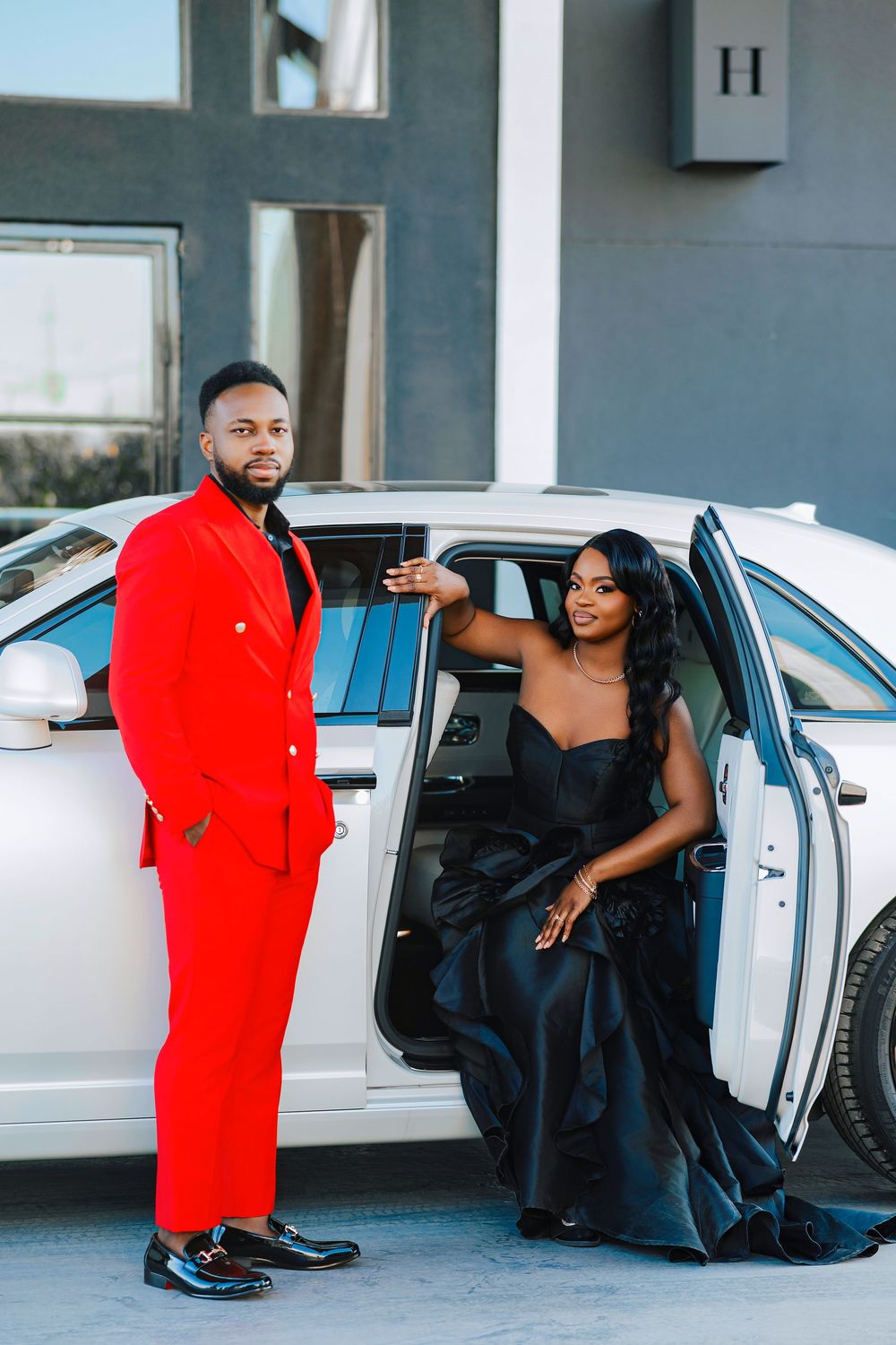 A couple in formal attire poses next to a car, with one person wearing a bright red suit and the other in black.