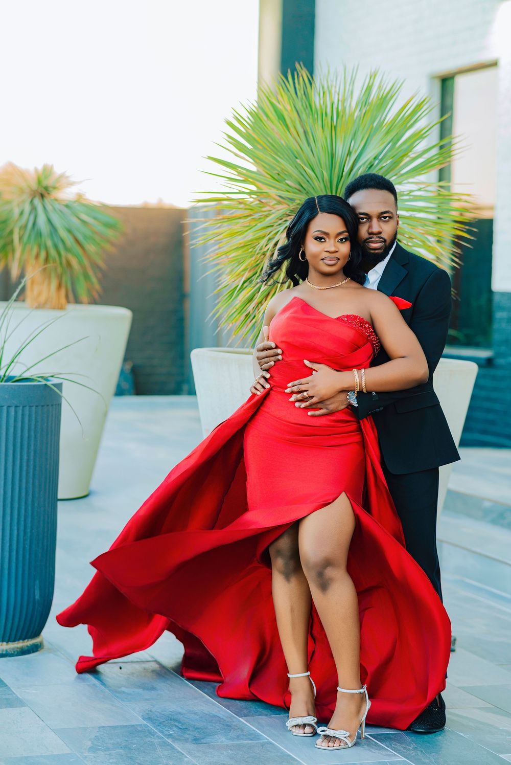 A couple poses outdoors against tropical plants, with the woman wearing a flowing red gown and the man in a black suit.