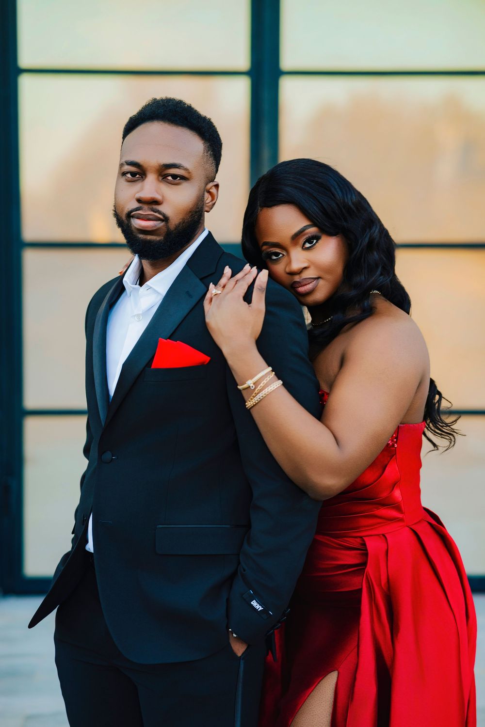 Elegant couple poses in formal attire with man in black suit and woman in flowing red gown against window backdrop.