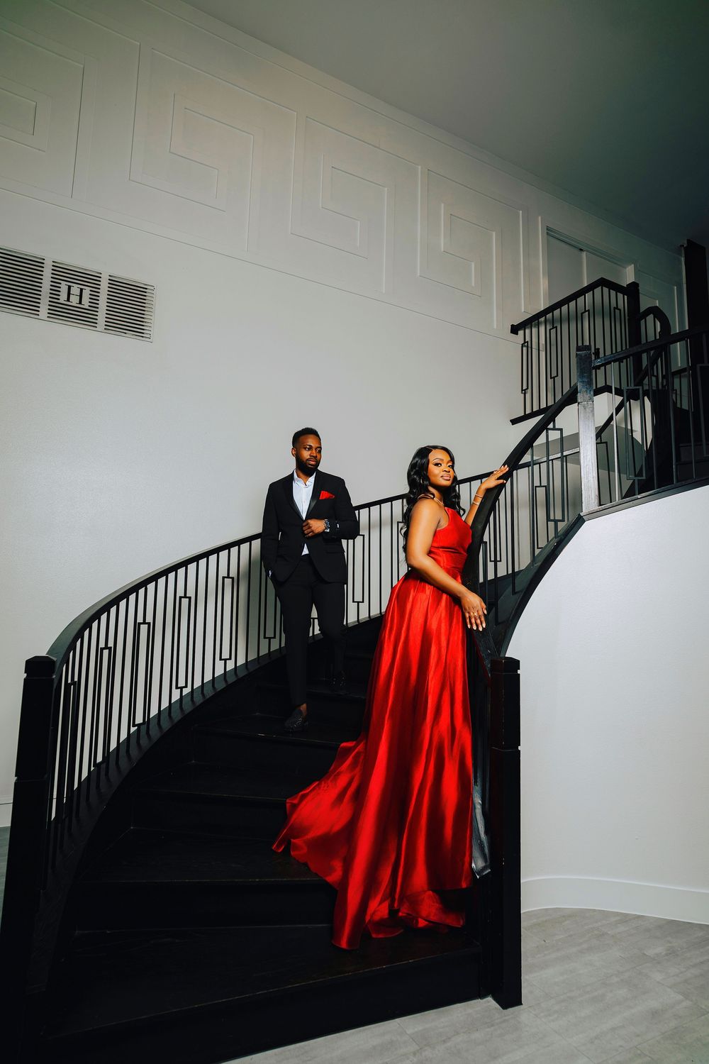 A couple in formal attire poses on a dramatic black staircase, with the woman wearing a flowing red evening gown.