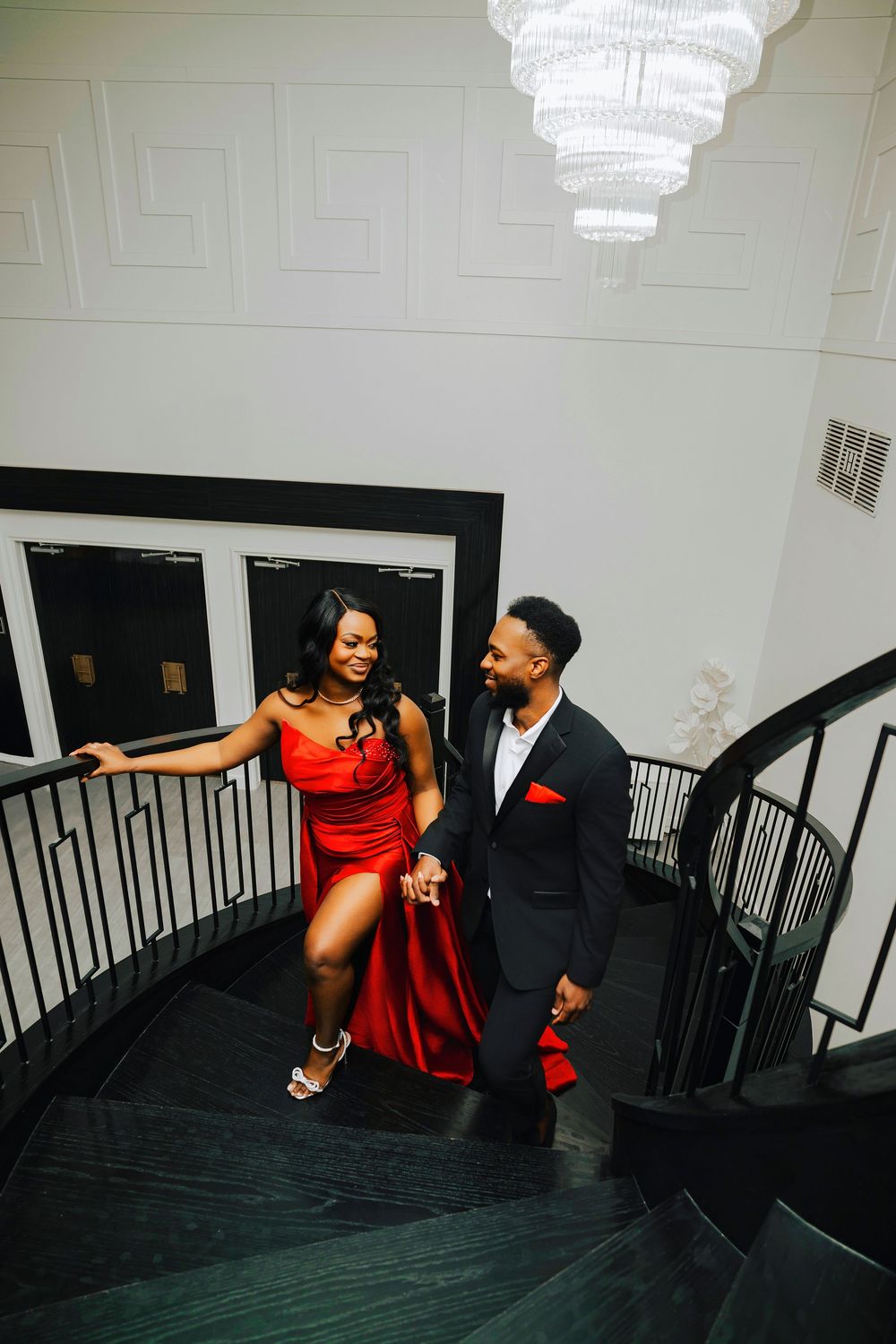 Romantic staircase scene with couple in formal attire, featuring red dress and black suit against white walls.