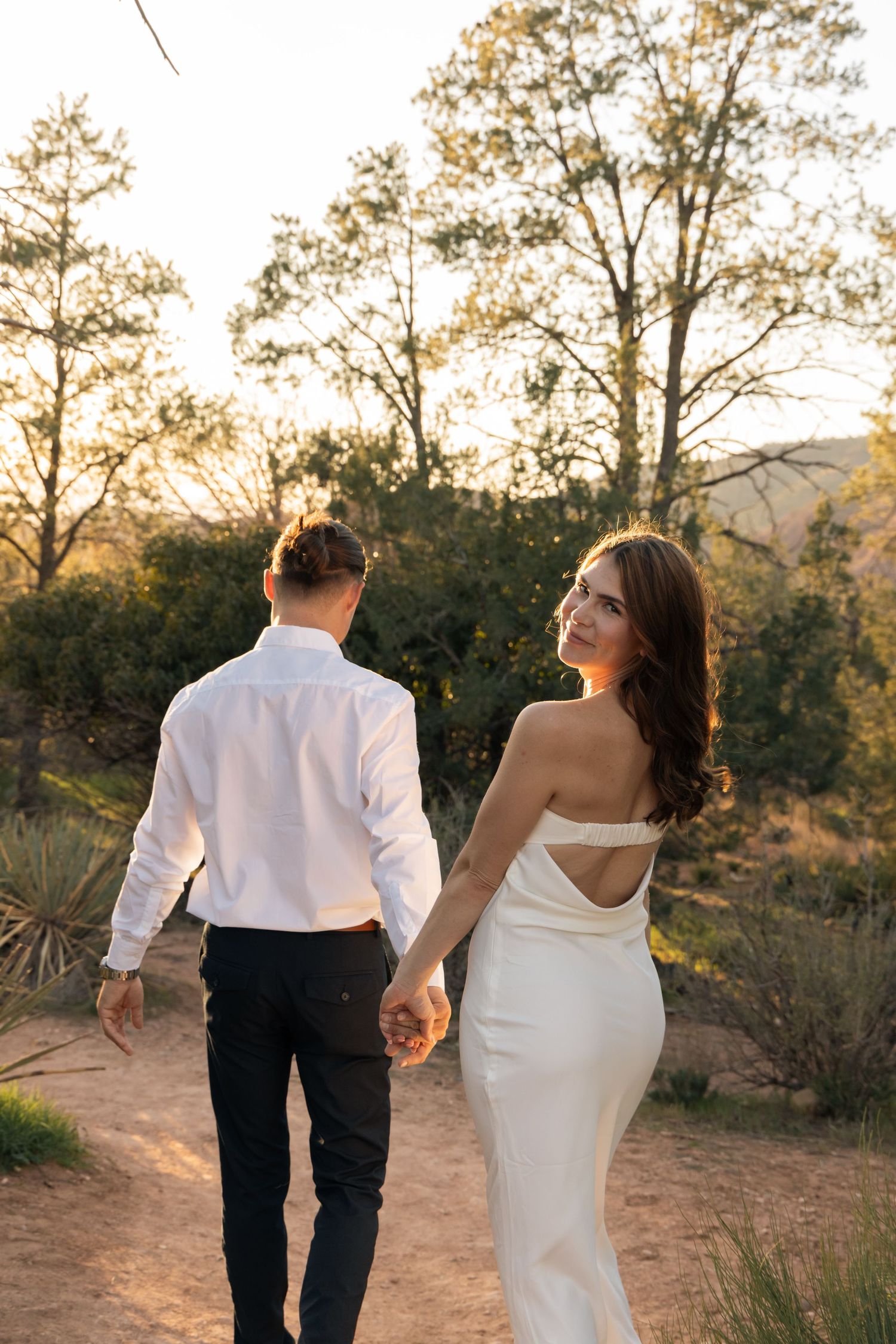 A couple in wedding attire walks hand in hand along a scenic mountain trail at sunset.