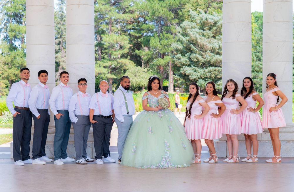 A quinceañera court with bridesmaids in pink dresses and groomsmen in grey suits posing by white columns.