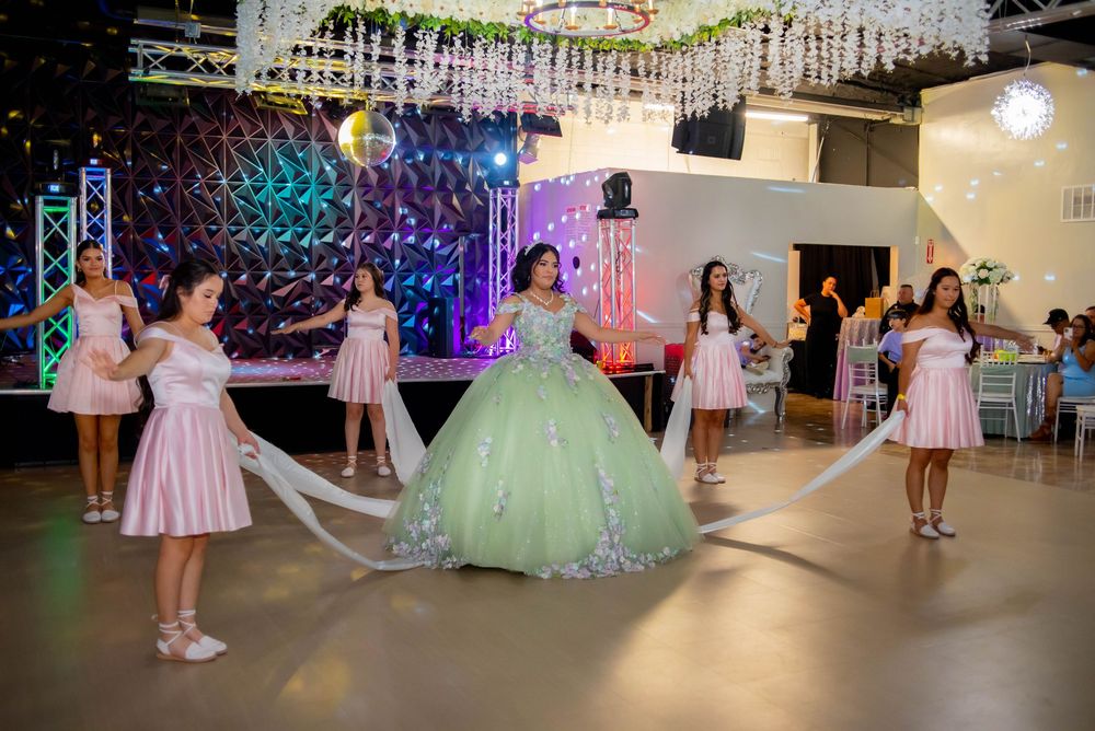 A festive quinceañera dance celebration with bridesmaids in pink dresses and decorative lighting.