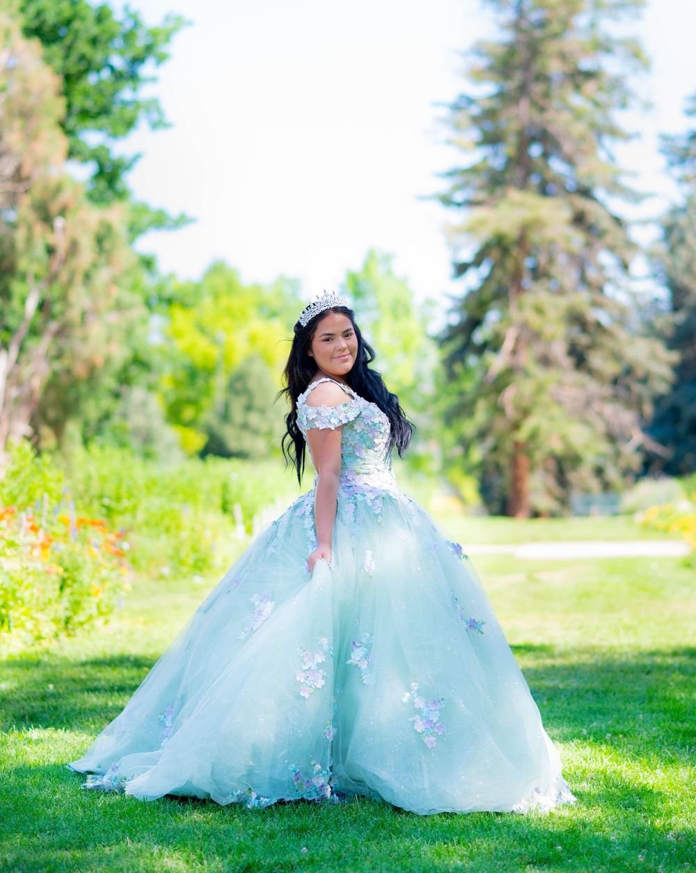 A stunning mint green quinceañera ball gown with floral appliques photographed in a sunny outdoor garden setting.