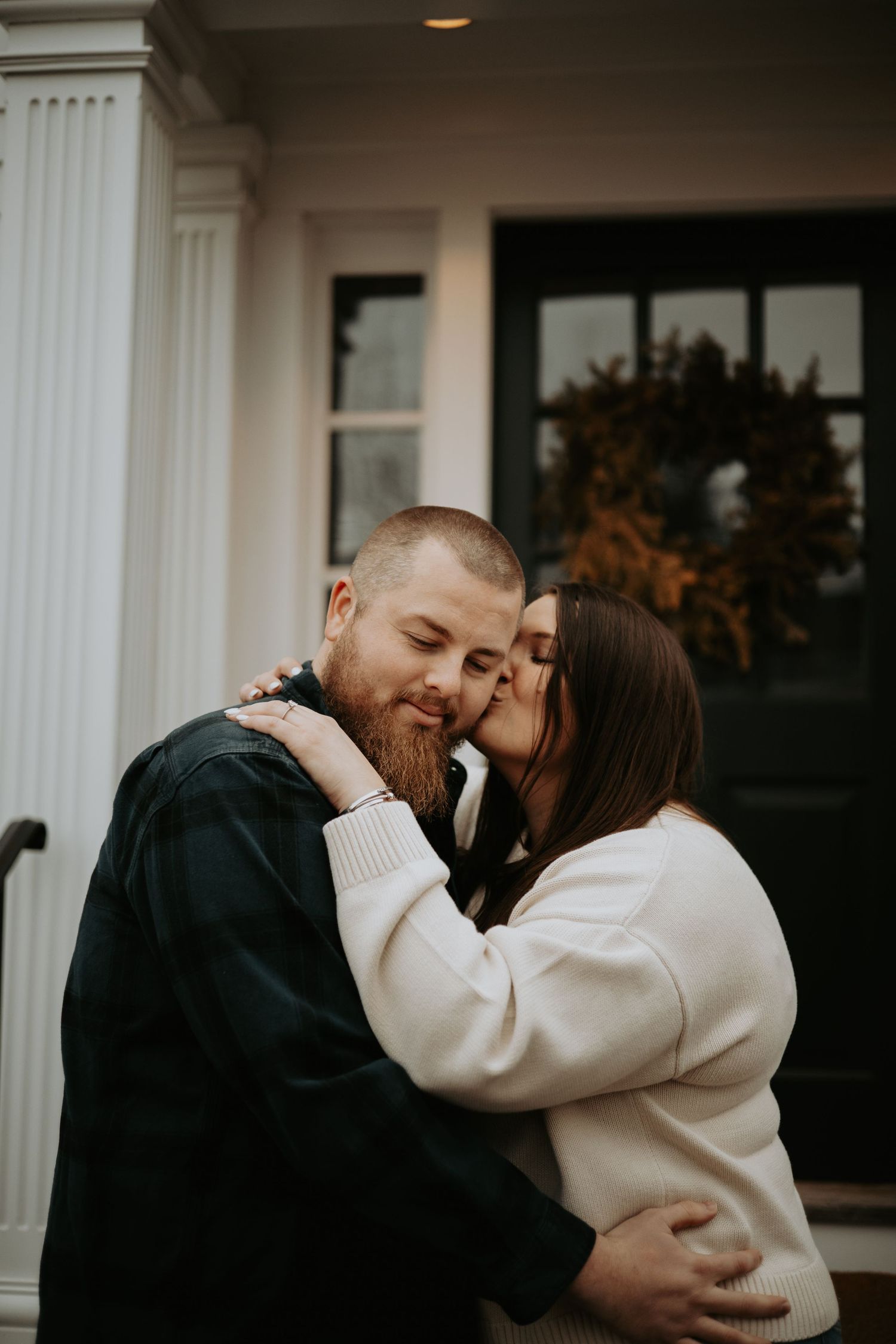 A romantic couple embraces outdoors in winter wearing black and cream colored sweaters against white pillars.