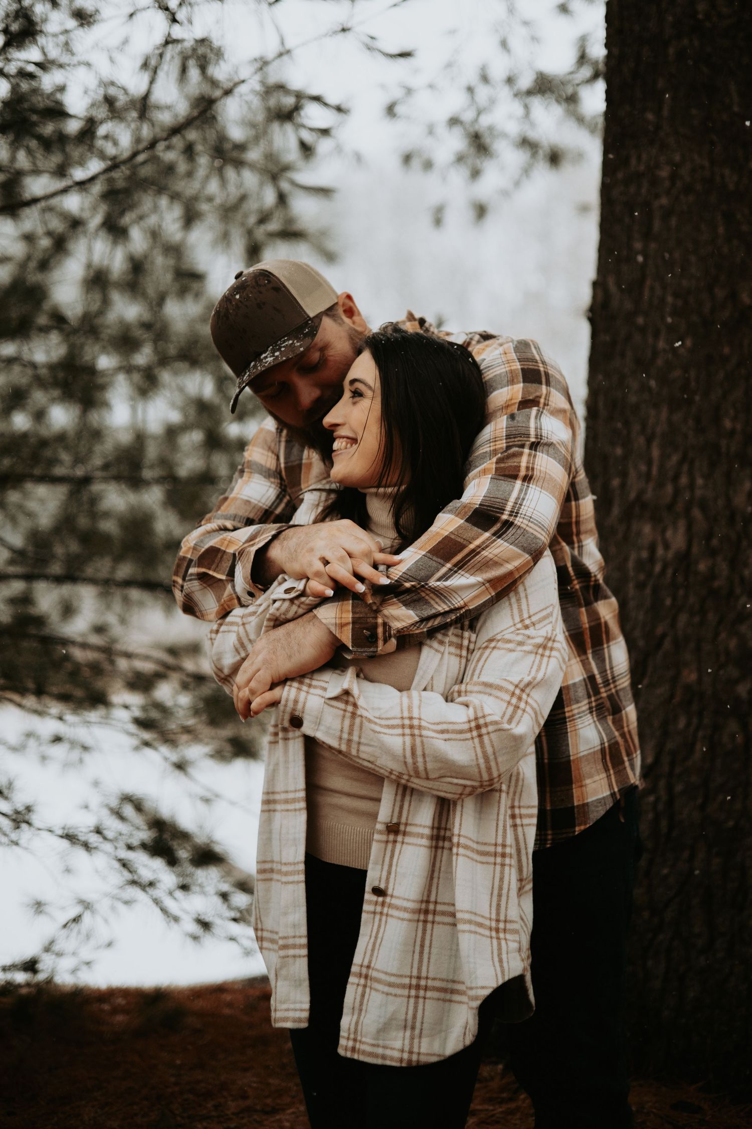 A couple embraces in a cozy plaid flannel shirt against a snowy winter forest backdrop.