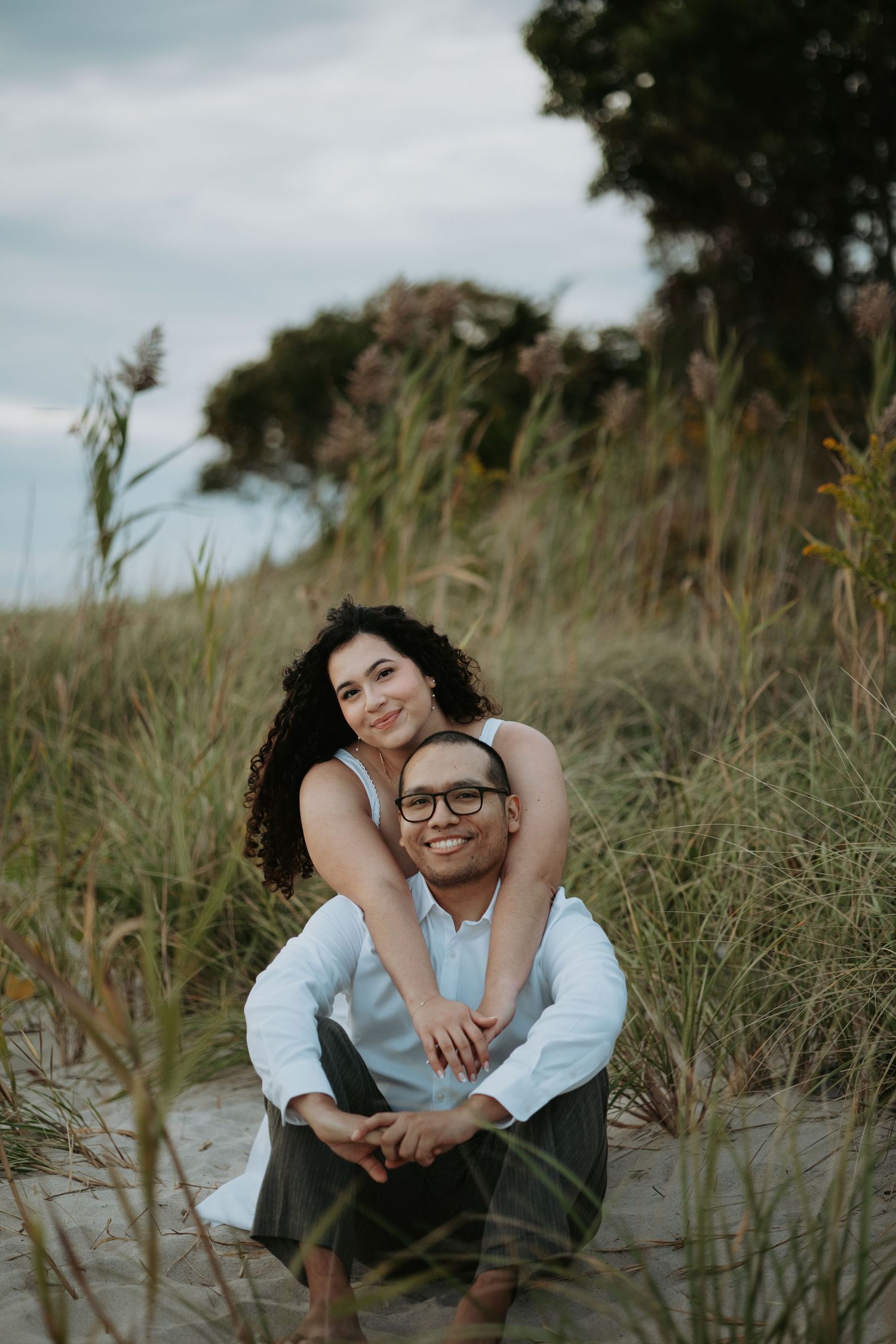 Smiling couple posing for an engagement photo on a sandy beach path, her arms wrapped around him, surrounded by tall dune grasses.