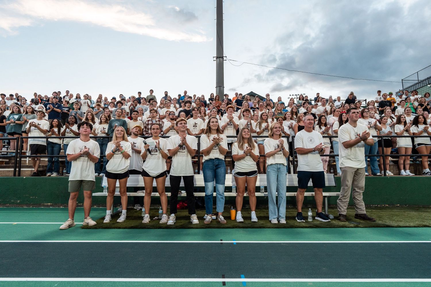 A panoramic view of a large crowd seated in white attire at an outdoor tennis tournament venue.