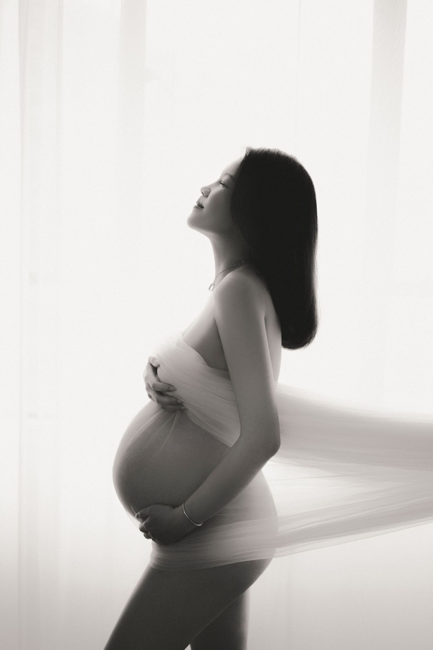 Elegant maternity photo in a light-filled room with vintage furniture, floral arrangements, and soft flowing curtains.