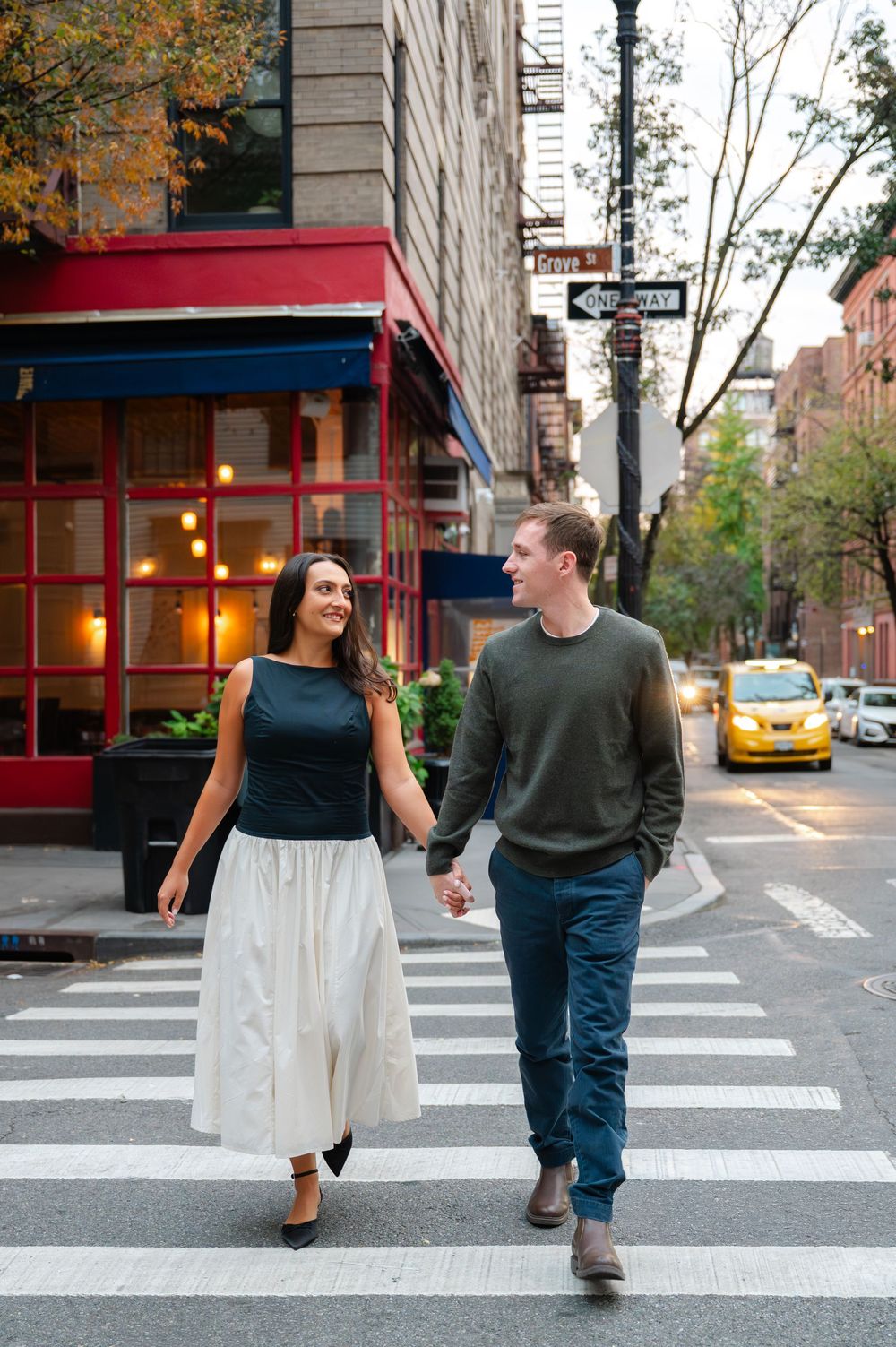 Couple walking across a crosswalk in New York City with fall foliage and storefronts in the background.