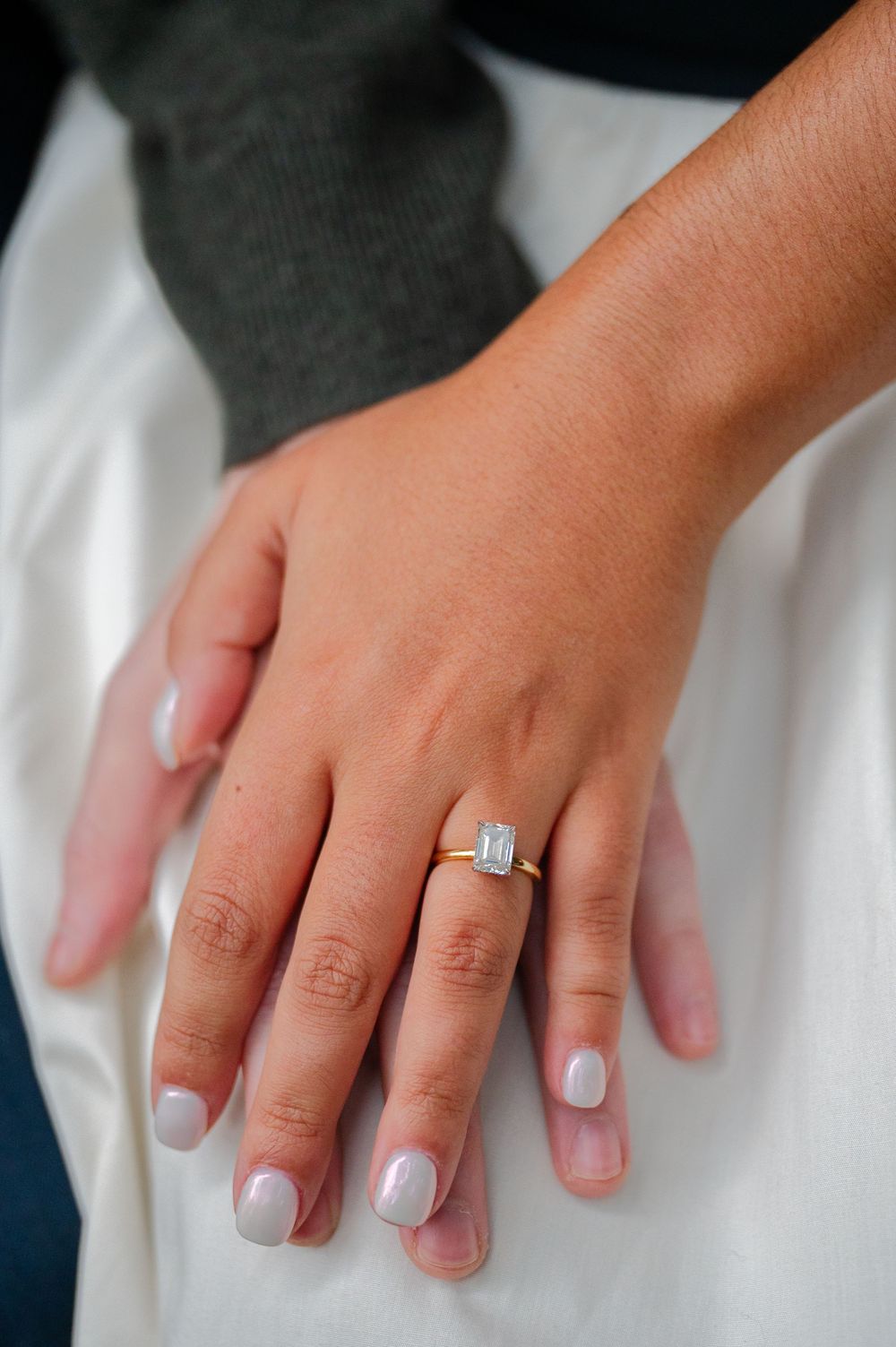 Close up of a hand wearing a white gold engagement ring with an emerald cut diamond on French manicured nails.
