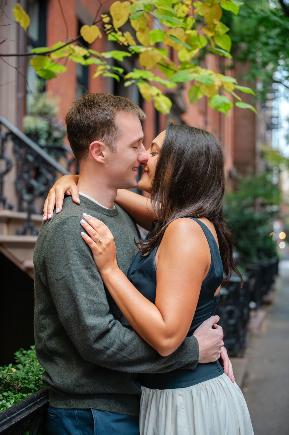 A romantic couple embraces on a tree-lined city street wearing grey and black outfits.