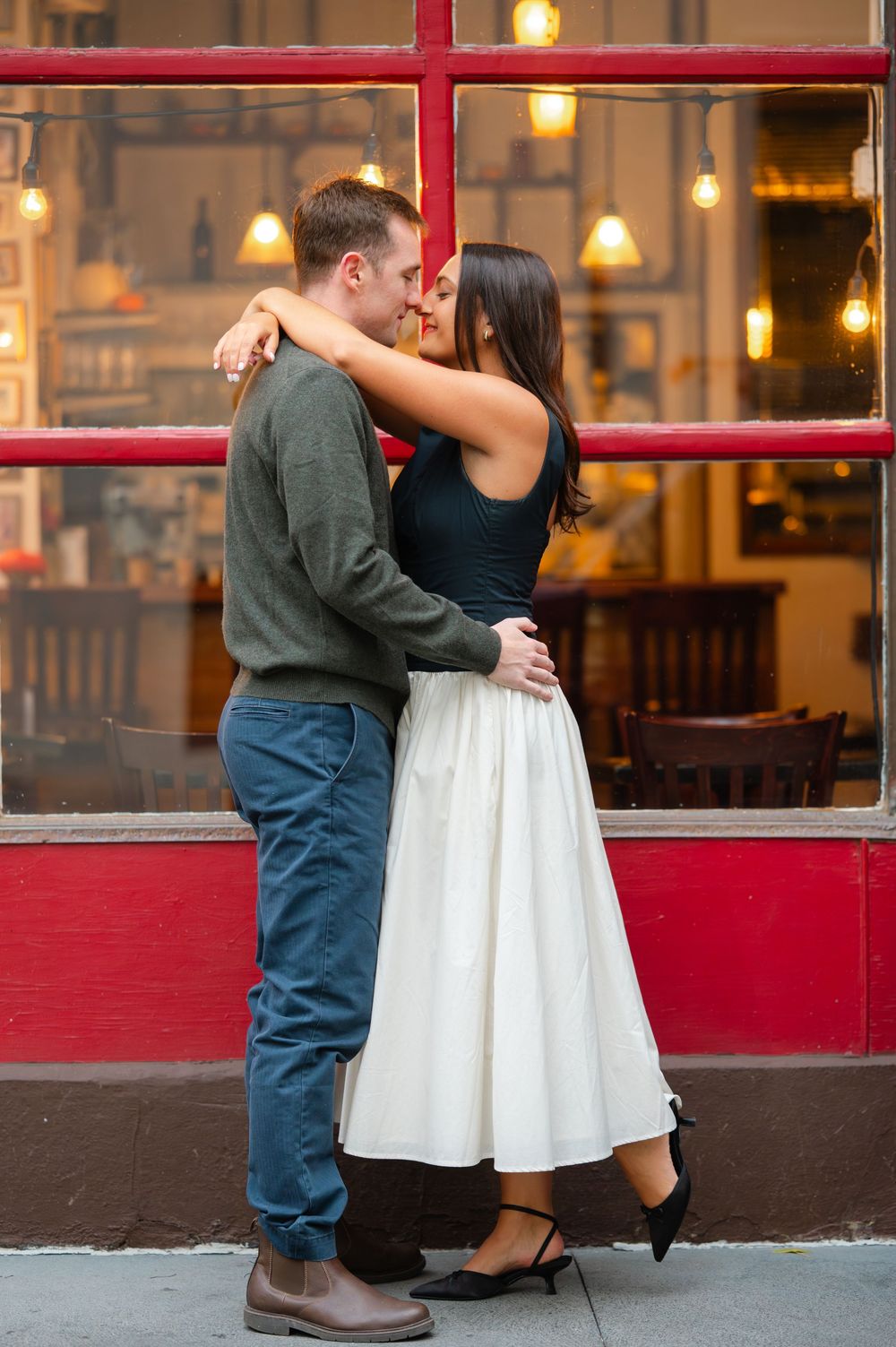 A couple embraces near a restaurant storefront with red trim and warm lighting in the evening.
