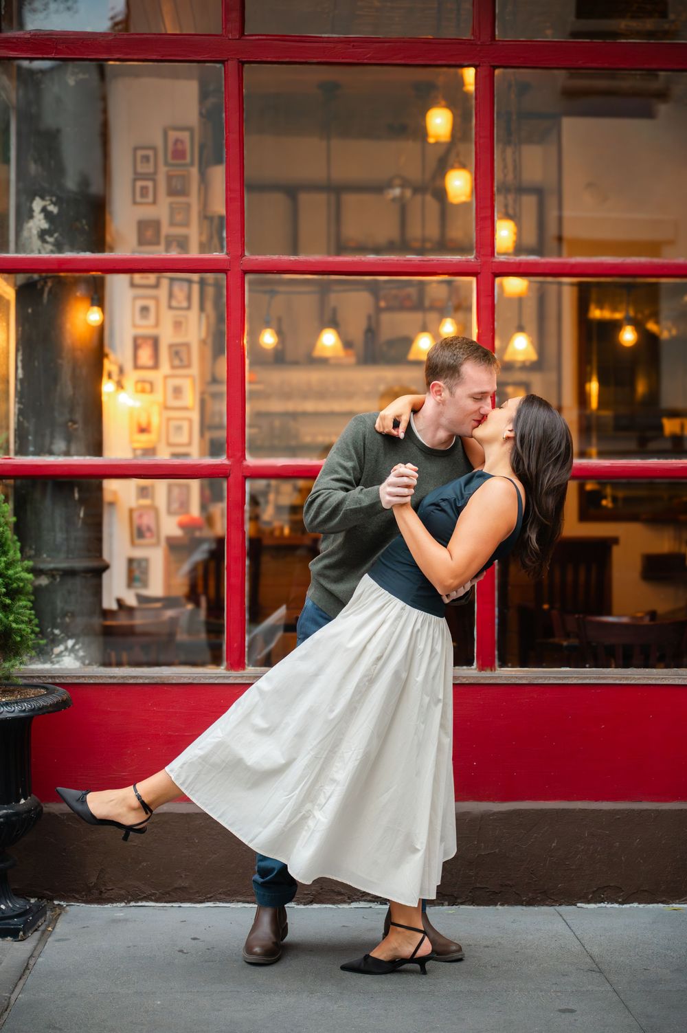 A man dip kisses his fiancee who is wearing a white and black dress with the red exterior of the Little Owl in the West Village behind them.
