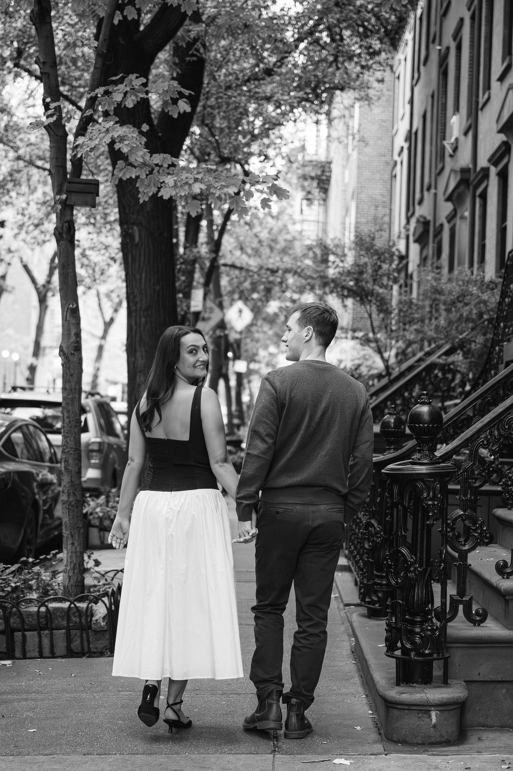 Couple walking hand in hand down a brownstone-lined street in black and white.