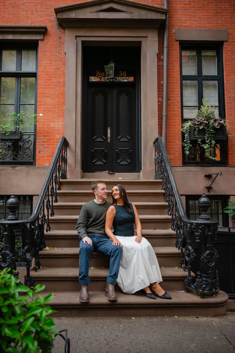 A couple sits together on brownstone steps in front of a brick building with black railings.