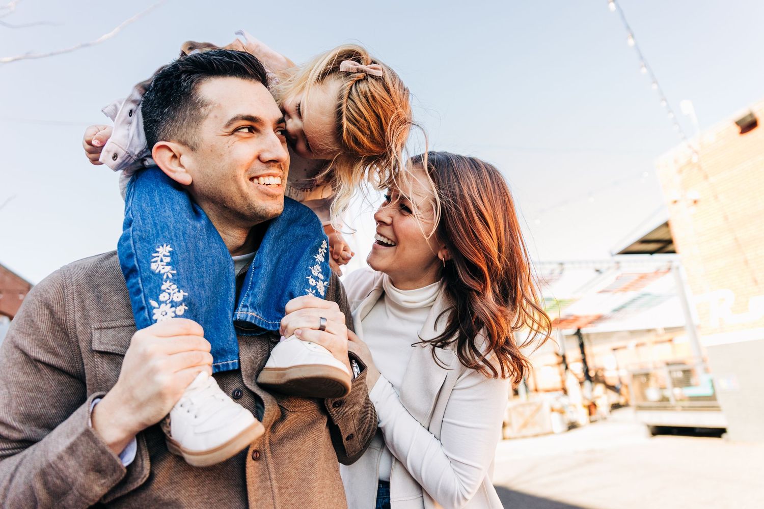 Couple laughing and sharing a tender moment with their toddler sitting on her dad's shoulders while walking together outdoors during sunset.