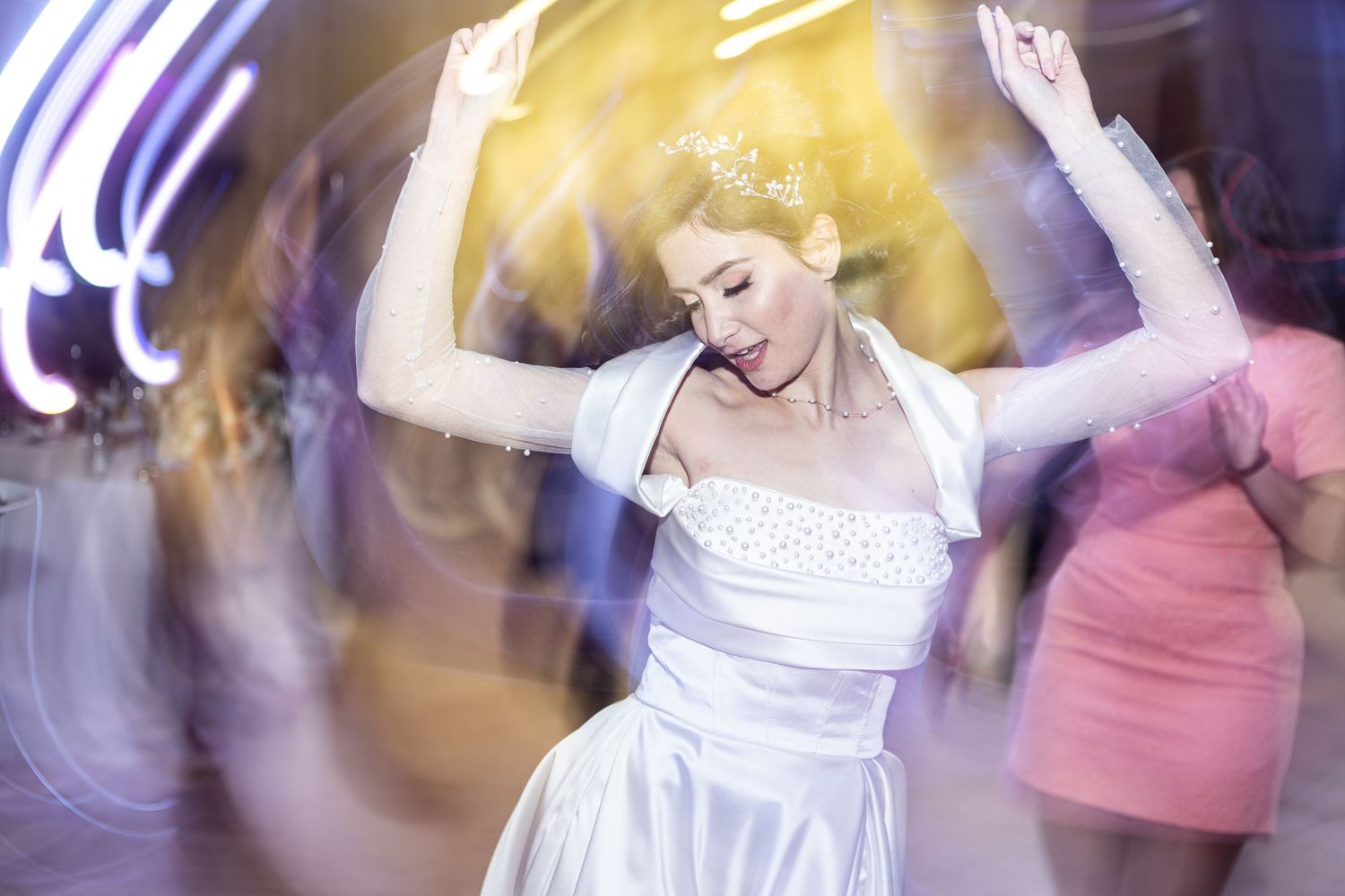 Bride in white strapless gown dances joyfully with arms raised at a vibrant wedding reception.