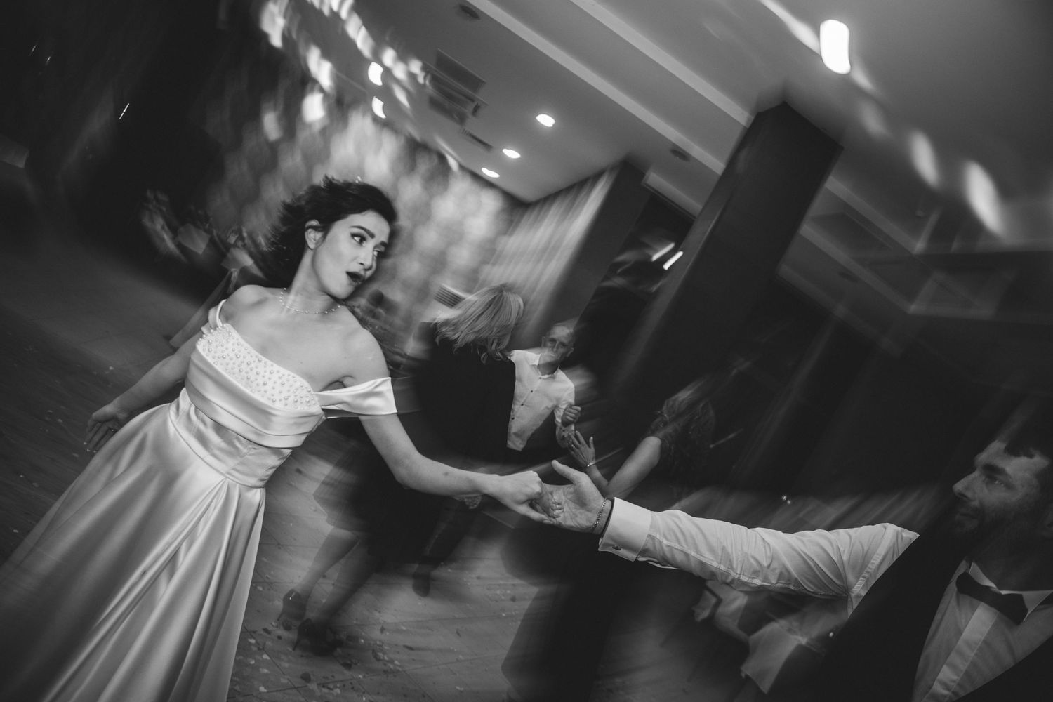 Bride in white off-shoulder gown dances elegantly with partner in black and white wedding reception photo.