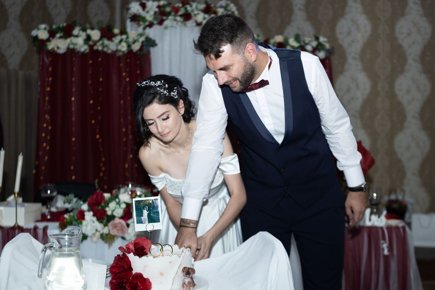 Bride and groom cut their white wedding cake decorated with red roses at elegant reception.