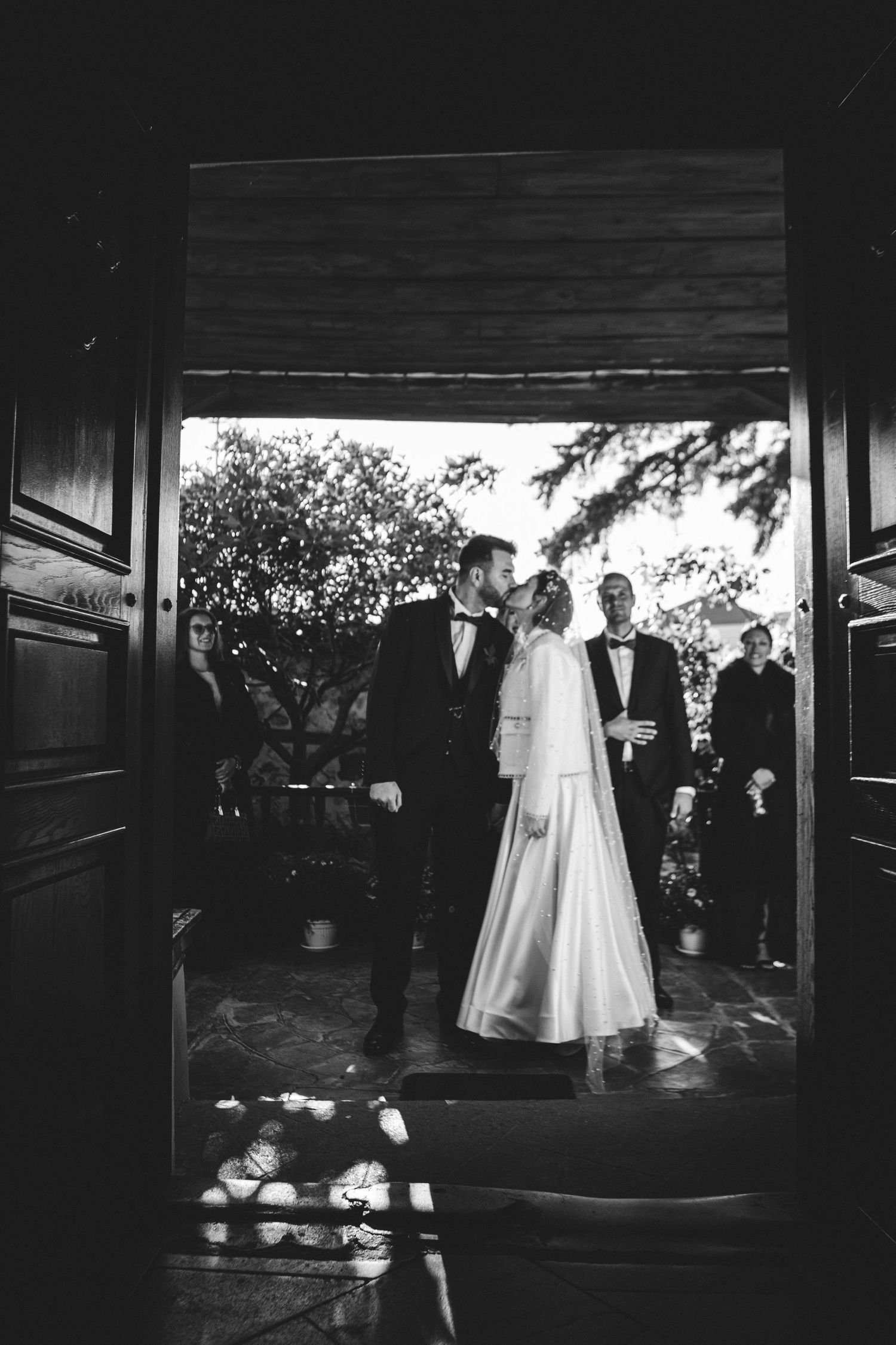 Black and white romantic shot of bride and groom kissing in doorway with guests behind.
