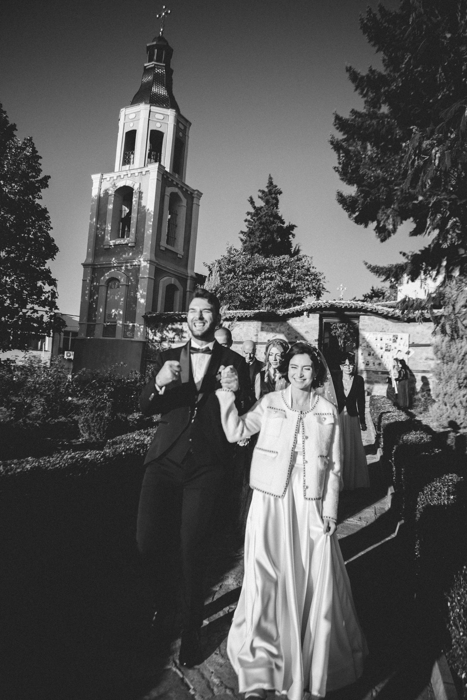 Black and white photo of joyful bride and groom walking outside historic church bell tower.