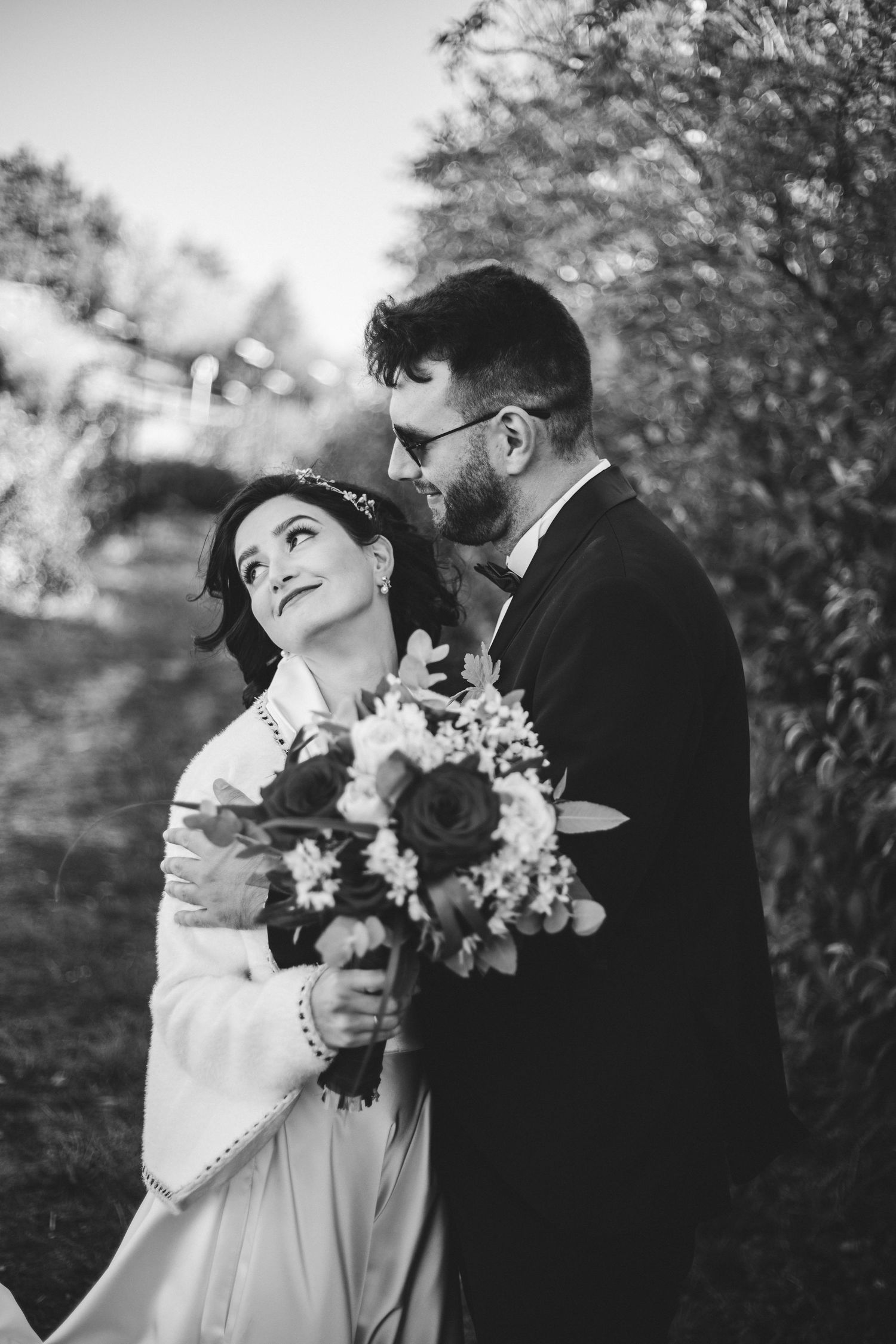 Black and white wedding photo of bride holding bouquet while groom embraces her from behind outdoors.