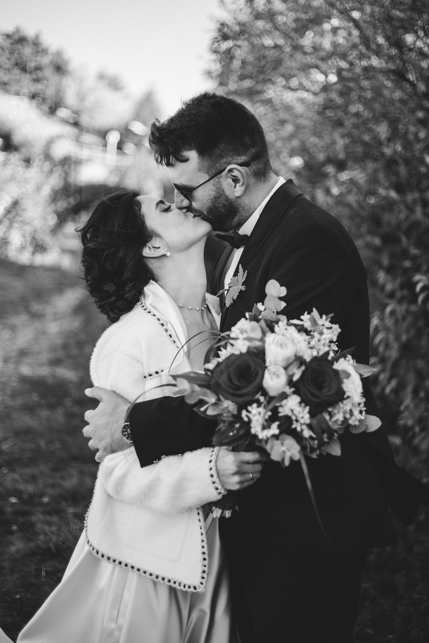 Black and white photo of bride and groom sharing a romantic kiss outdoors while holding wedding bouquet.