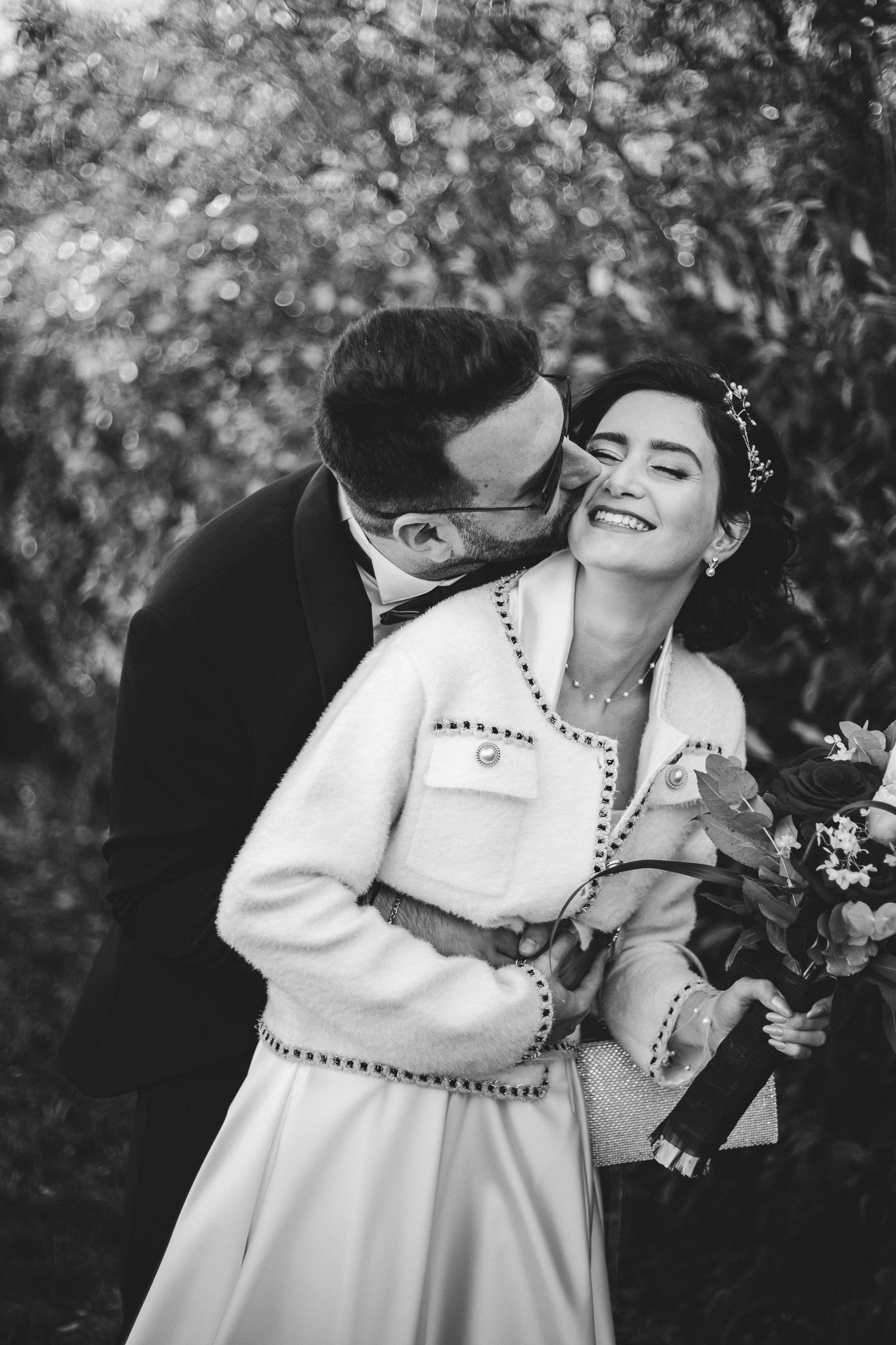 Groom kissing bride on cheek as she laughs joyfully holding floral bouquet in black and white photo.