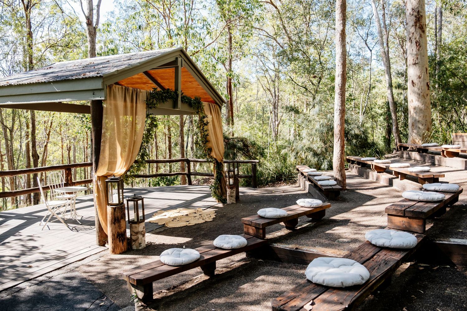 Rustic wooden picnic shelters with benches line a shaded path in a peaceful forest setting.