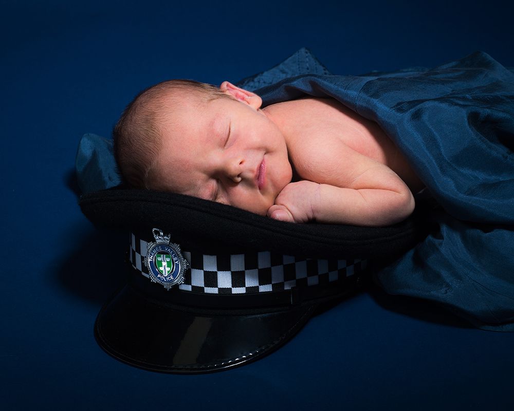 Newborn baby sleeping peacefully on a dark blue police hat against a deep blue backdrop. Photography by Abigail's Studio, High Wycombe.