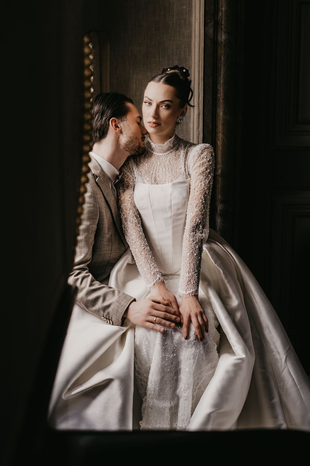 A romantic wedding portrait in moody lighting shows a couple embracing in elegant white lace and satin attire.