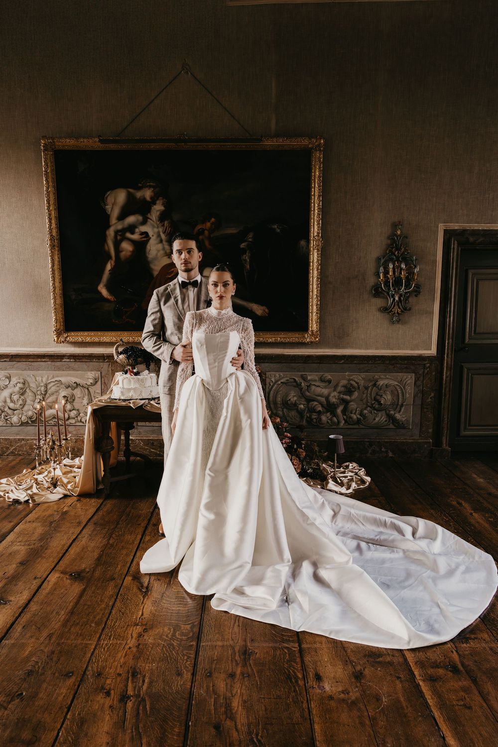 Elegant couple poses in formal wedding attire inside historic room with ornate decor and wooden floors.