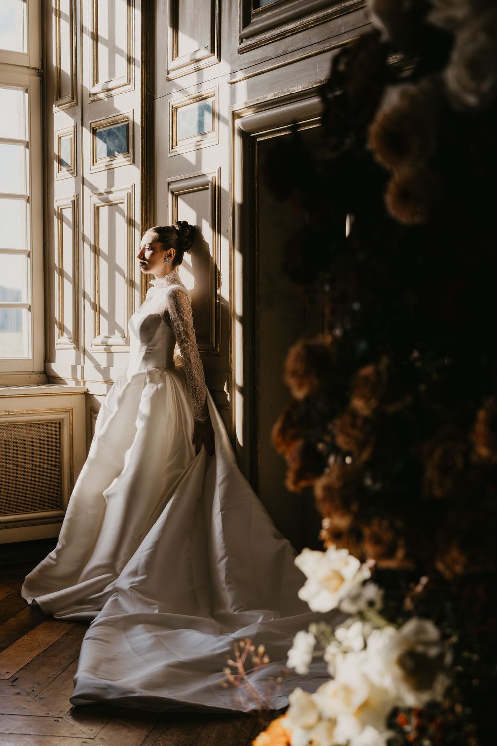 A bride in an elegant white gown stands by a sunlit window, with white flowers decorating the ornate interior space.