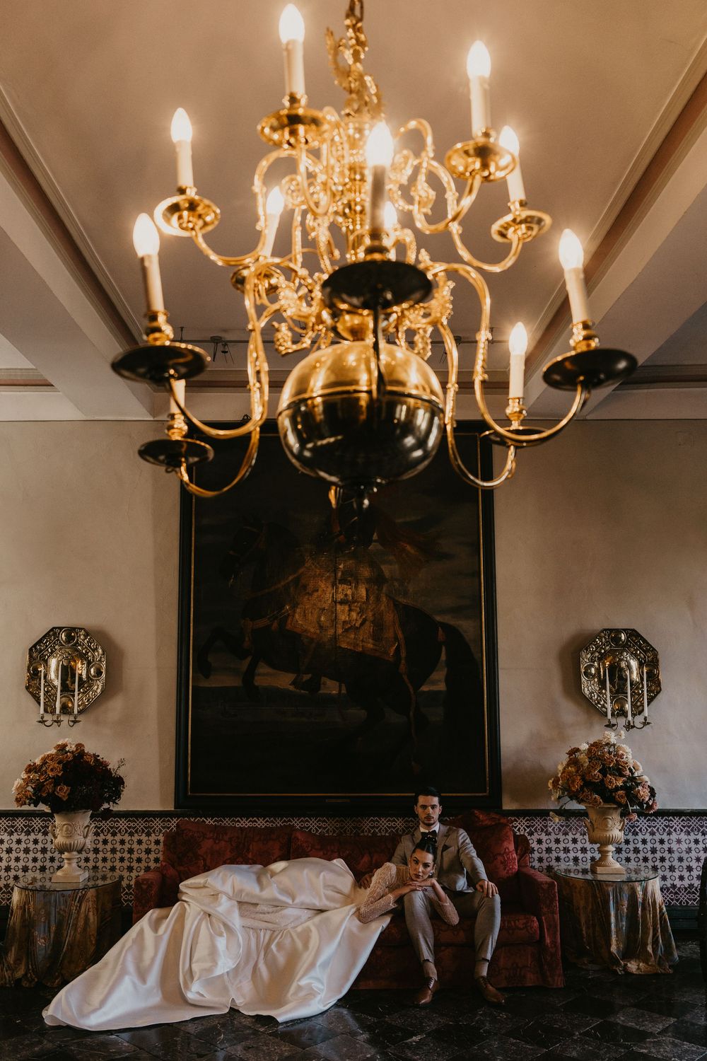 Elegant brass chandeliers with glowing candle lights illuminate an ornate dining room with classic decor and dark walls.