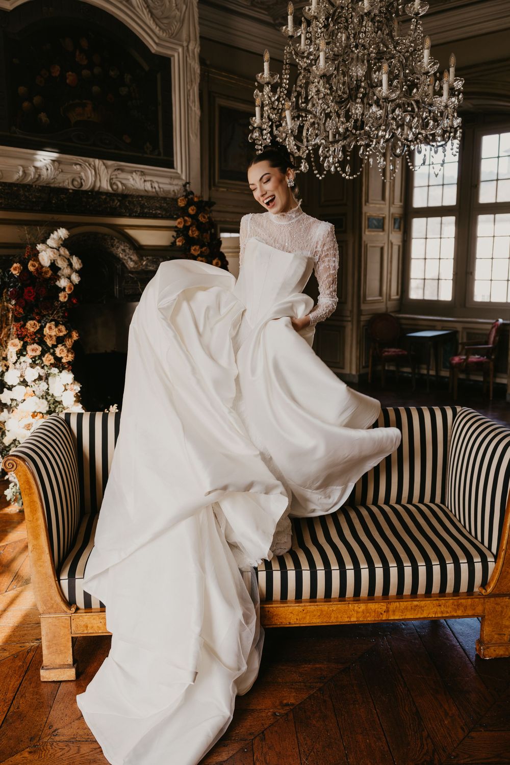 A bride in a flowing white wedding dress twirls playfully on a black and white striped sofa in an elegant room.